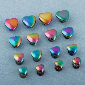Puede incluir: Una colección de tiradores de armario iridiscentes en forma de corazón. Los tiradores muestran un espectro de colores, incluyendo oro, rosa, azul y verde. Están dispuestos sobre una superficie texturizada de color azul claro. Estos tiradores decorativos son ideales para muebles.