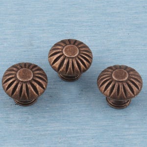 Könnte beinhalten: Drei bronzefarbene Schrankknöpfe mit floralem Design. Jeder Knopf hat eine kreisförmige Oberfläche mit radialen Rillen und einem zentralen, erhabenen Kreis. Die Knöpfe sind vor einem hellblauen Hintergrund angeordnet.