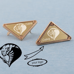 Peut inclure: Deux épingles triangulaires dorées avec un motif de cobra et le mot "Cobra" gravé. Les épingles ont une bordure en or rose et sont posées sur un fond bleu clair. Un logo de cobra et les mots "your logo" sont également présents.