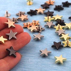 May include: A collection of star-shaped metal rivets in various colors, including gold, silver, bronze, and black. Some rivets are held in a hand, while others are scattered on a light blue surface. The rivets have a screw-in post on the back.