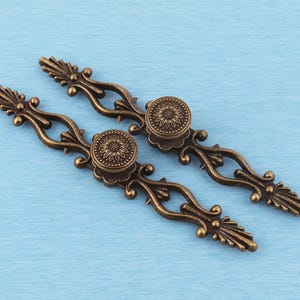 May include: Two antique bronze-coloured metal drawer pulls featuring an elongated, openwork scroll design with a central circular knob. Each knob is decorated with a detailed sunflower-like pattern, and the ends of the pulls are shaped like pointed, ribbed leaves.
