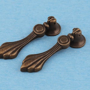 Puede incluir: Dos tiradores de cajón de metal en tono bronce antiguo con diseño colgante. Cada tirador consta de una base de montaje circular conectada a un mango articulado y alargado que se ensancha en forma de abanico con tres nervaduras verticales.