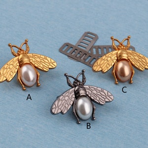 May include: Three bee-shaped brooches with pearl accents. The brooches are gold, silver, and black.