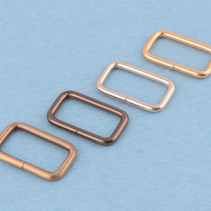 May include: Four rectangular metal loops arranged diagonally. Each loop features a distinct metallic finish, including shades of copper, bronze, and silver. The rectangular hardware components have rounded corners and a smooth, polished surface texture.
