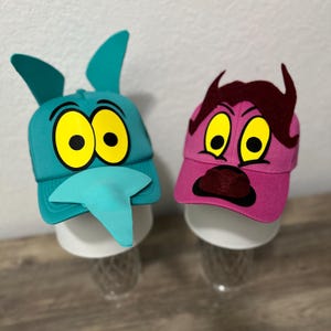 May include: Two cartoon character baseball caps. One is teal with large yellow eyes, a blue tongue, and tall ears. The other is pink with yellow eyes and maroon hair and a surprised expression.