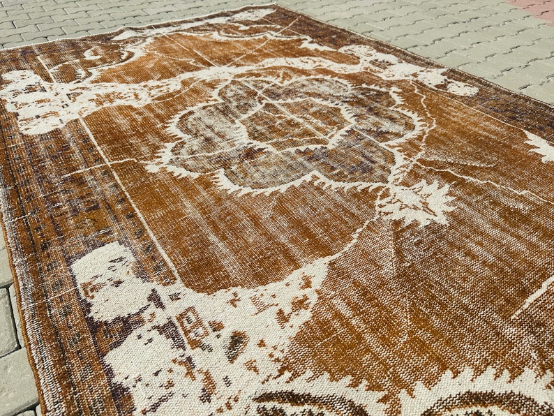 Puede incluir: Una alfombra vintage con un aspecto descolorido y desgastado. La alfombra tiene un fondo marr&oacute;n con un patr&oacute;n floral blanco. La alfombra est&aacute; hecha de lana y tiene una textura tejida a mano.