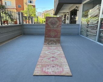 Puede incluir: Una alfombra larga y estrecha con un patrón geométrico rosa y beige. La alfombra está doblada por la mitad y se encuentra sobre una superficie gris.
