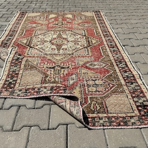Op de afbeelding: Een vintage Turks tapijt in rood, bruin en crème. Het tapijt heeft een geometrisch patroon met een centraal medaillon. Het tapijt is dubbelgevouwen en ligt op een grijs betonnen oppervlak.