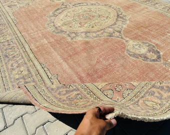 May include: A large, rectangular, vintage rug with a distressed, faded appearance. The rug features a central medallion design in shades of beige, brown, and burgundy, set against a muted coral background. The border has a detailed pattern.