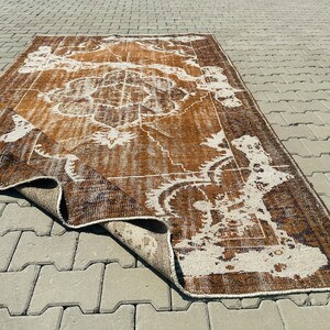 Puede incluir: Una alfombra vintage marr&oacute;n y blanca con un patr&oacute;n floral. La alfombra est&aacute; doblada por la mitad y se encuentra sobre una superficie de hormig&oacute;n gris.