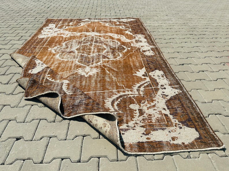 Puede incluir: Una alfombra vintage marr&oacute;n y blanca con un patr&oacute;n floral. La alfombra est&aacute; doblada por la mitad y se encuentra sobre una superficie de hormig&oacute;n gris.