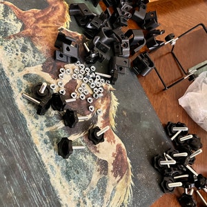 May include: A close-up of a variety of black plastic hardware pieces, including clamps, knobs, and nuts. The hardware is scattered on a table with a brown wooden surface. The hardware is arranged in a random pattern.