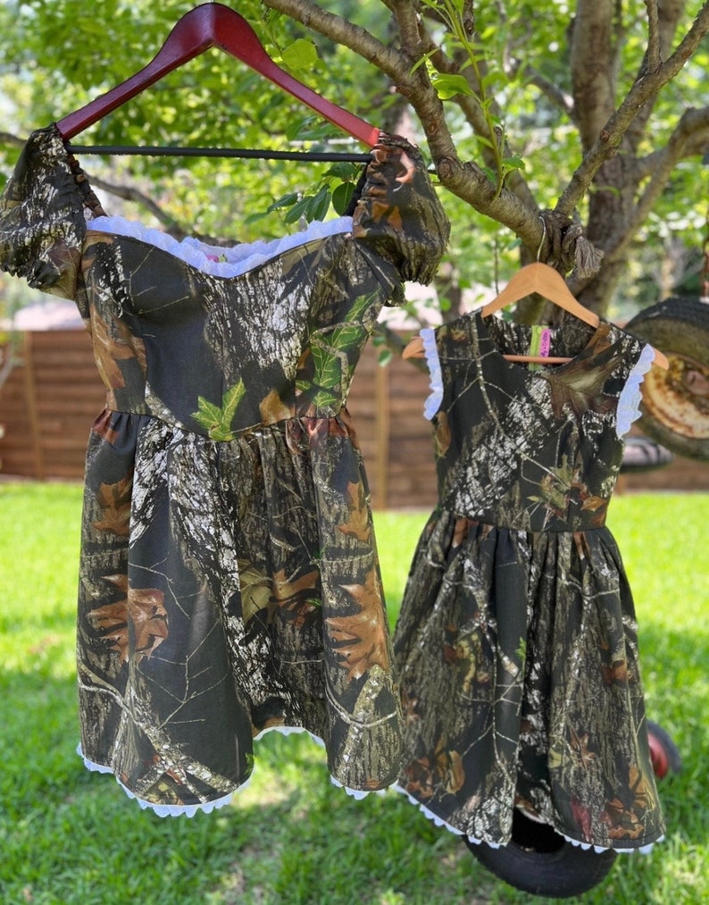 Puede incluir: Dos vestidos de estampado de camuflaje con ribete de encaje blanco. El vestido m&aacute;s grande tiene mangas cortas y cuello redondo. El vestido m&aacute;s peque&ntilde;o tiene tirantes sin mangas y cuello redondo.