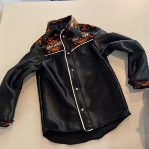 Może przedstawiać: Długa koszula satynowa w kolorze czarnym z białym obszyciem i wzorem płomieni. Koszula zawiera logo Harley-Davidson w kolorze pomarańczowym i białym.