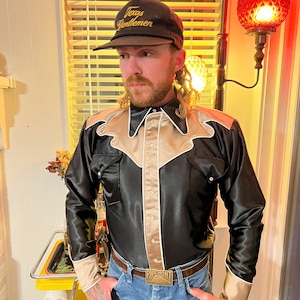May include: A man wearing a black satin western shirt with beige trim and a brown baseball cap that says "Texas Gentlemen". He is also wearing blue jeans and a belt with a silver buckle that says "Philmont".