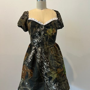 Puede incluir: Un vestido corto de camuflaje verde, marr&oacute;n y blanco con un ribete de encaje blanco alrededor del escote y el dobladillo. El vestido tiene mangas cortas y abullonadas.