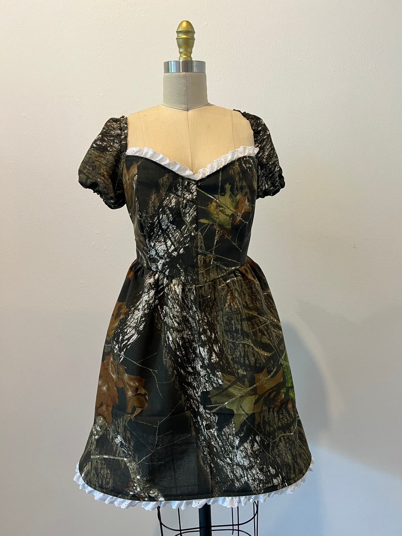 Puede incluir: Un vestido corto de camuflaje verde, marr&oacute;n y blanco con un ribete de encaje blanco alrededor del escote y el dobladillo. El vestido tiene mangas cortas y abullonadas.