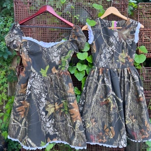 Puede incluir: Dos vestidos de estampado de camuflaje con ribete de encaje blanco. Los vestidos tienen mangas cortas y est&aacute;n colgados en perchas de madera.