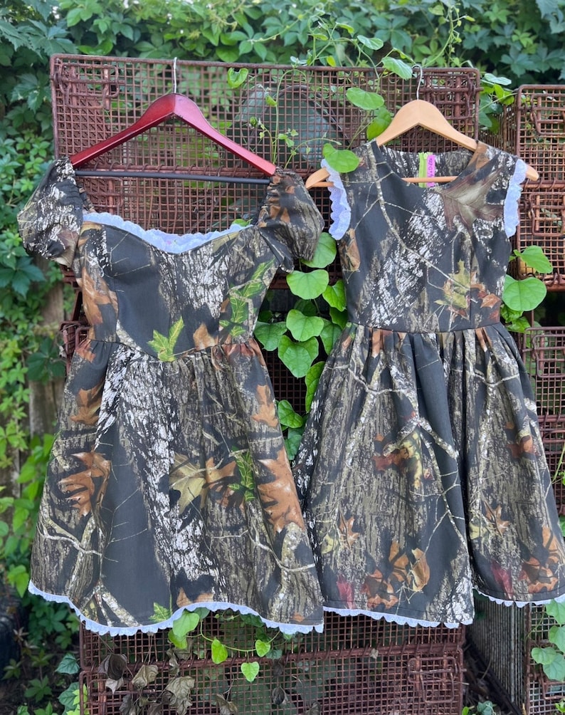 Puede incluir: Dos vestidos de estampado de camuflaje con ribete de encaje blanco. Los vestidos tienen mangas cortas y est&aacute;n colgados en perchas de madera.