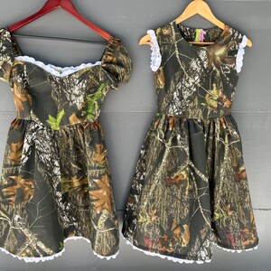 Puede incluir: Dos vestidos con estampado de camuflaje y ribete de encaje blanco. Los vestidos son sin mangas y tienen un corpi&ntilde;o ajustado con una falda completa.