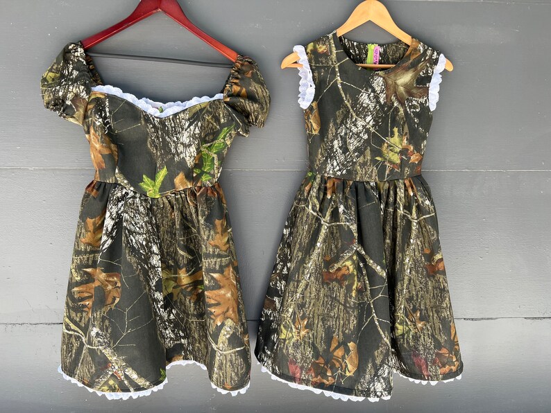 Puede incluir: Dos vestidos con estampado de camuflaje y ribete de encaje blanco. Los vestidos son sin mangas y tienen un corpi&ntilde;o ajustado con una falda completa.