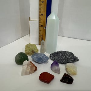 以下が含まれることがあります： 高さ約10cmの木製定規の横に立つ淡い青白色のクリスタルポイントと、緑、青、赤、紫、黒の色合いの小さなタンブルストーンや原石がいくつか含まれる、さまざまな鉱物標本のコレクション。