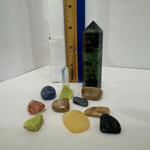 Puede incluir: Una colección de varias piedras preciosas y cristales pulidos, incluida una torre hexagonal de color verde oscuro, junto a una regla de madera que muestra una altura de unos 10 cm. El surtido presenta piedras en tonos amarillos, azules, verdes y marrones.