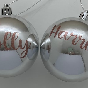 May include: Two silver Christmas baubles with rose gold lettering. The baubles are personalised with the names "Molly" and "Harriet".