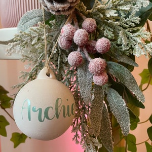 Può includere: Un addobbo natalizio rotondo e bianco con il nome "Archie" scritto in corsivo verde chiaro. L'addobbo è appeso a un ramo di verde con bacche rosse e una pigna.