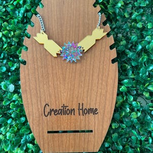 May include: A gold-colored necklace with a starburst pendant, displayed on a wooden jewelry stand. The necklace has a chain with a white and silver pattern. The stand has the words "Creation Home" engraved on it.