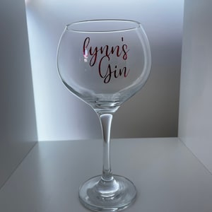 Könnte beinhalten: Ein klares Glas-Gin-Glas mit einem rosa Vinyl-Aufkleber, auf dem "lynn's Gin" steht.