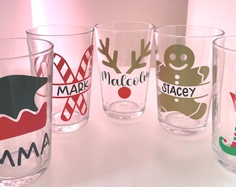 May include: Five clear glass cups with Christmas-themed designs and names. The designs include a candy cane, a reindeer, a gingerbread man, an elf, and a Santa hat. The names on the cups are Mark, Malcolm, Stacey, Amy, and Emma.