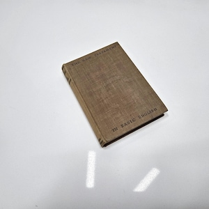 May include: A hardcover book titled "The New Testament in Basic English" with a tan cover. The book is rectangular and lies on a white surface. The text is printed in black.