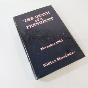 May include: A hardcover book titled "THE DEATH of a PRESIDENT" with the author's name, William Manchester, and the date November 1963 printed in pink on a dark blue cover. The book is slightly angled, showing its cover and spine.