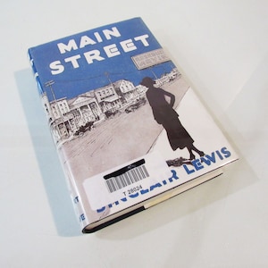 May include: A hardcover book titled "Main Street" by Sinclair Lewis. The cover features a blue sky background with white text and an illustration of a town street scene and a silhouetted figure. A barcode label is affixed to the book.