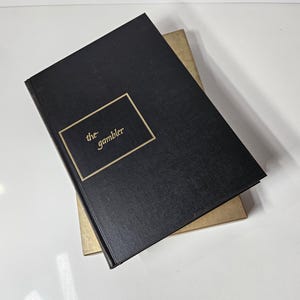 May include: A black book titled "the gambler" with gold lettering and a gold border. The book is on top of a tan book. The books are on a white surface.