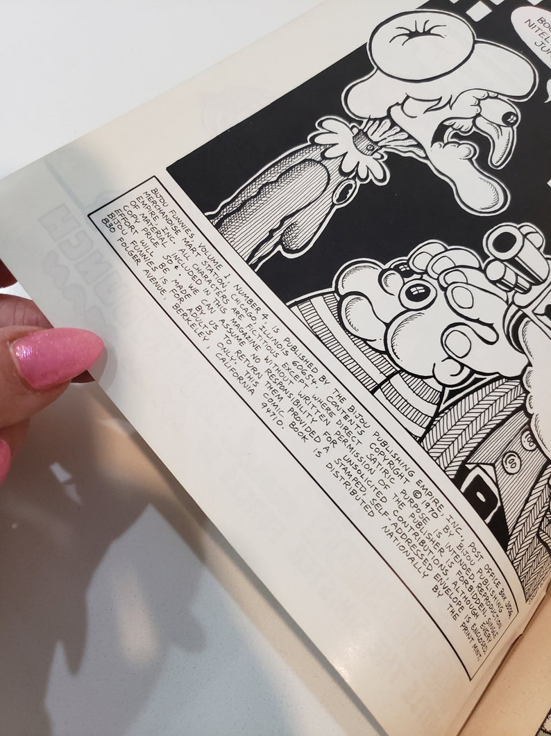 May include: A black and white comic book illustration with cartoon characters. The open comic book displays artwork and text, including the title "BIJOU FUNNIES" and publishing details. The image is held by a hand with pink nail polish.