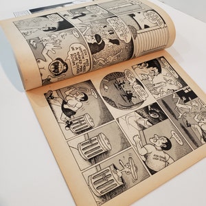 May include: A comic book open to a double-page spread with black and white cartoon illustrations. The panels depict various scenes with speech bubbles. Text includes "EEK! MISS HINTON! THERE'S A NAKED MAN IN OUR LOCKER ROOM!" The pages have a yellowed hue.