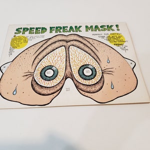 May include: A vintage "Speed Freak Mask" paper mask with a cartoonish design. The mask features two large, bloodshot eyes with blue irises and a light brown skin tone. Text on the mask reads "Perfect for Parties!" and "Teens!"