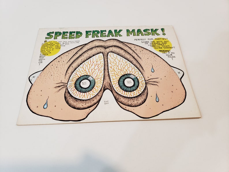 May include: A vintage "Speed Freak Mask" paper mask with a cartoonish design. The mask features two large, bloodshot eyes with blue irises and a light brown skin tone. Text on the mask reads "Perfect for Parties!" and "Teens!"