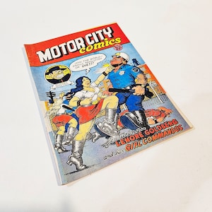 May include: A vintage comic book titled "MOTOR CITY COMICS" with a colorful illustration of a woman in a red skirt and boots being confronted by a police officer. The comic book cover features the text "JOIN THE WORLD...OR DIE!!!" and "Starring LENORE GOLDBERG and her GIRL COMMANDOS."
