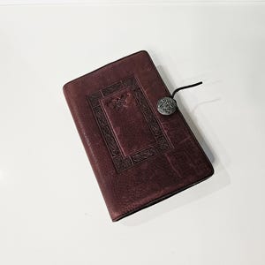 May include: A burgundy leather journal with an embossed Celtic knot design and a decorative metal clasp. The journal is rectangular and closed with a black cord and a silver-toned button clasp. The cover has a textured appearance.
