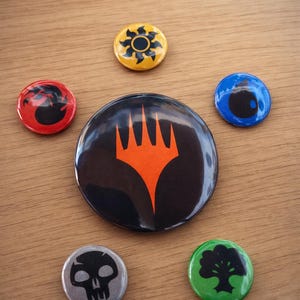 May include: A selection of circular, brightly coloured badges displaying different graphic designs. The badges feature symbols in red, yellow, blue, green, and silver, with a large black badge in the centre with an orange symbol. They are arranged on a wooden surface.