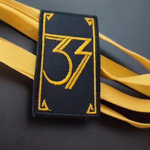 Può includere: Una toppa rettangolare nera con il numero "33" ricamato in oro. La toppa ha un bordo e dettagli angolari dorati. Un nastro giallo è drappeggiato attorno alla toppa su uno sfondo scuro.