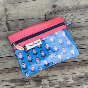 May include: A clear zippered pouch with a pink top and a blue patterned design. The pattern features a repeating illustration of a woman with glasses. The pouch has a black zipper and a small tag that reads "JDStudios480".