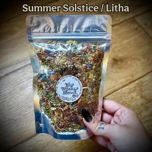May include: A transparent, resealable bag filled with dried herbs and flowers. The bag has the text "Summer Solstice / Litha" at the top. A circular sticker with the text "The Witches Hovel" is on the front. The bag is held by a hand.