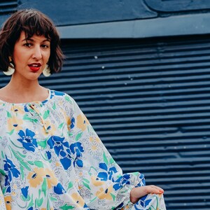 Pode incluir: Uma mulher vestindo um vestido branco com um padrão floral azul e amarelo. O vestido tem mangas compridas e uma cintura franzida. A mulher está em pé em frente a uma parede azul.