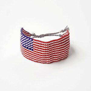 May include: A red, white, and blue beaded bracelet with a woven American flag design. The bracelet has gray string ties.
