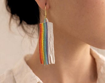 Puede incluir: Un pendiente dorado con un diseño rectangular con cuentas. El pendiente presenta un arcoíris de colores, incluyendo rojo, naranja, amarillo, verde, azul y morado, con flecos de cuentas blancas. El pendiente se lleva en la oreja de una persona.