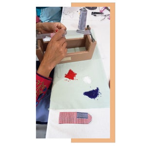 May include: A person is making a beaded American flag. The flag is red, white, and blue and is being made on a loom. There are piles of red, white, and blue beads next to the loom.